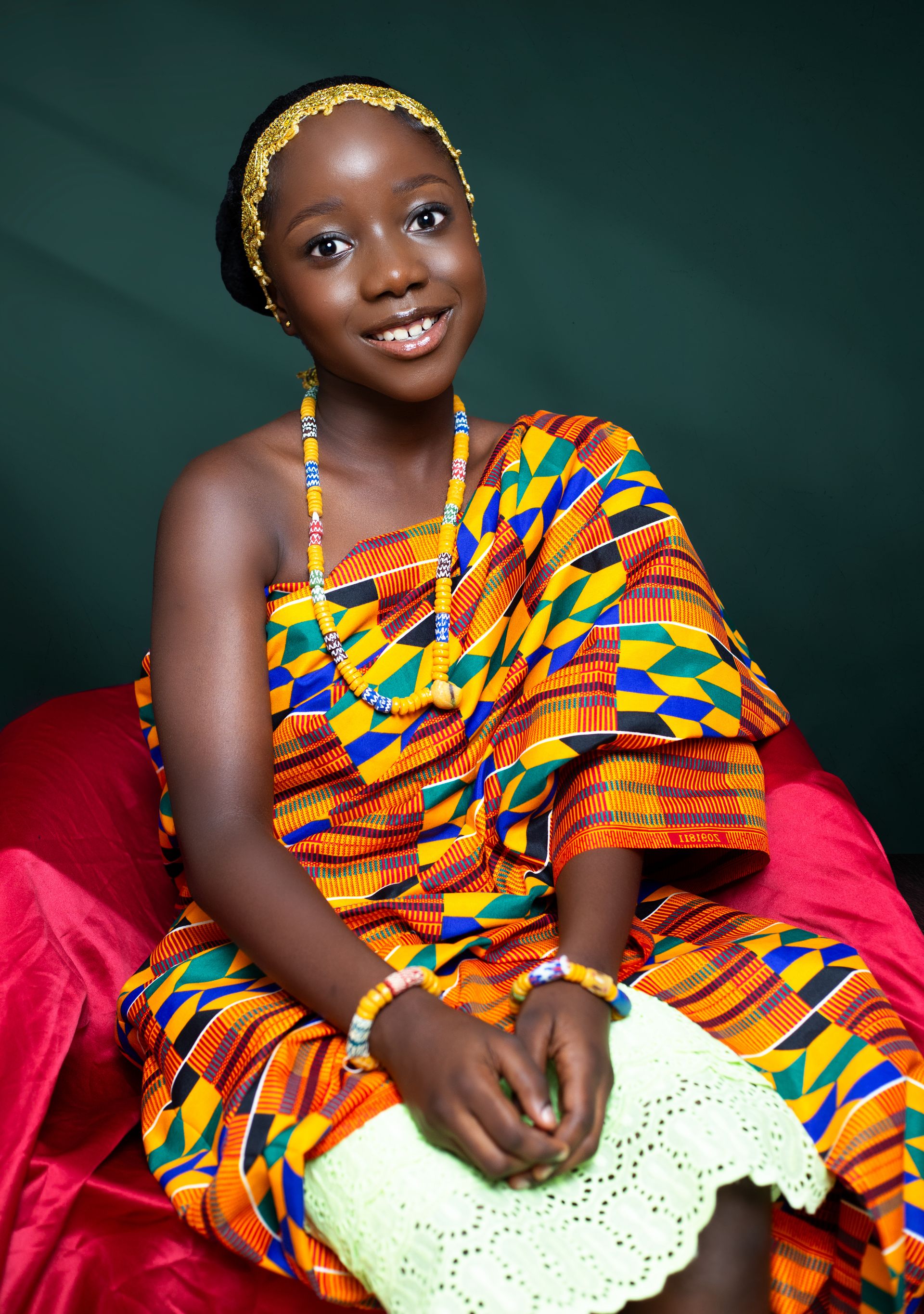 Portrait of a young woman seated in a vivid kente-print outfit holding a woven fan